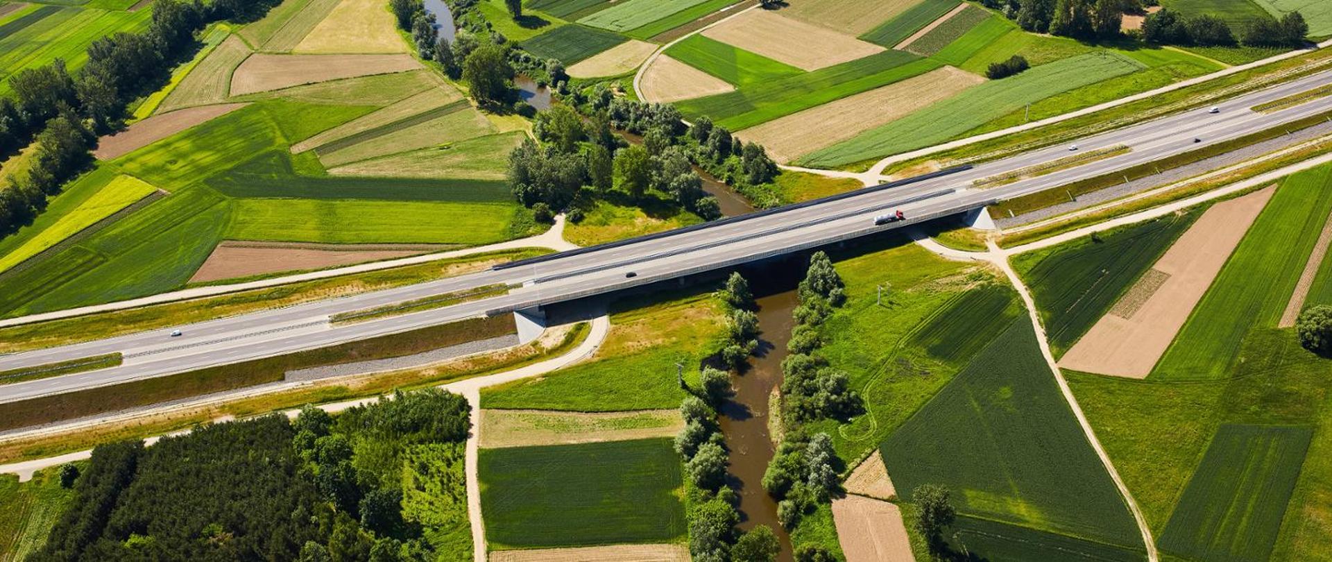 Na zdjęciu widoczna jest droga, most nad rzeką oraz otaczające infrastrukturę pola.