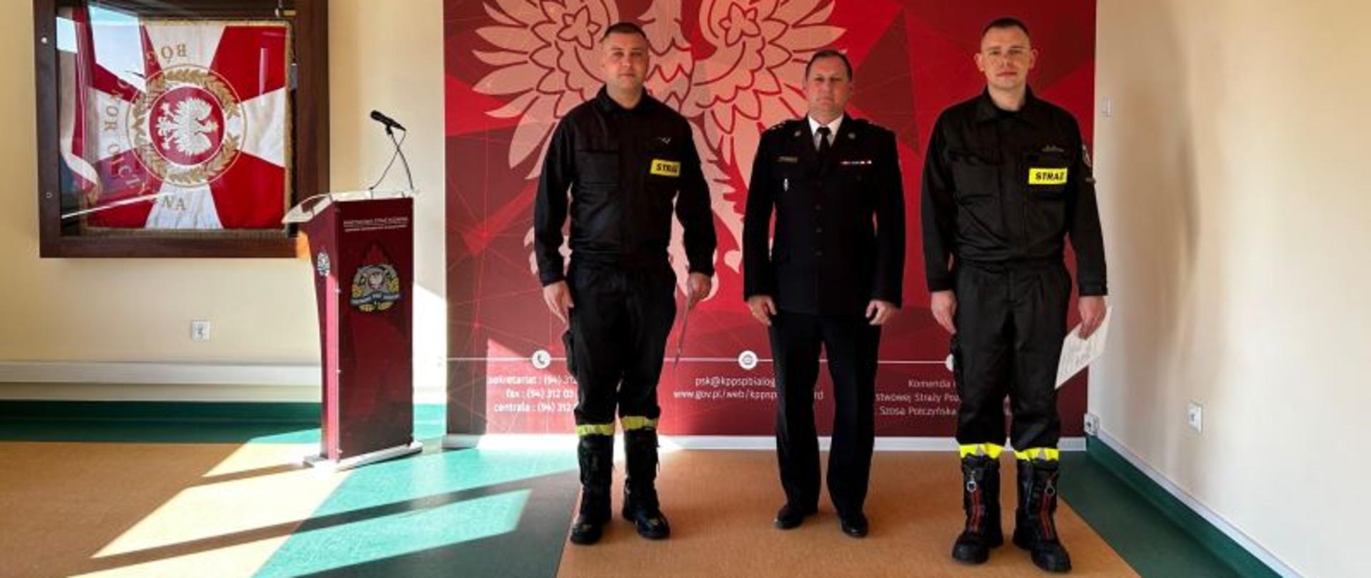 Przed ścianką informacyjną KP PSP Białogard stoi trzech strażaków. Dwaj w ubraniach koszarowych, jeden w ubraniu wyjściowym. Ścianka z godłem Polski w kolorze czerwonym.