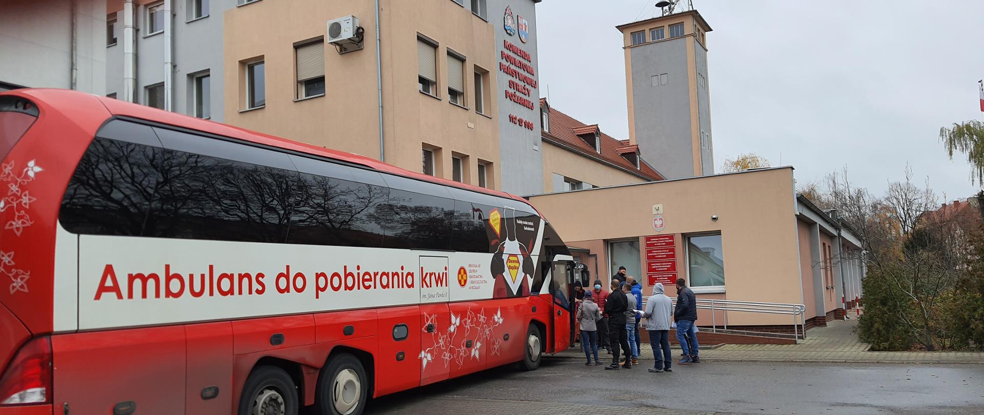 Honorowi krwiodawcy. Autobus Regionalnego Centrum Krwiodawstwa i Krwiolecznictwa w Poznaniu zaparkowany przed budynkiem Komendy Powiatowej PSP w Kościanie. Osoby stoją w kolejce do oddania krwi. 
