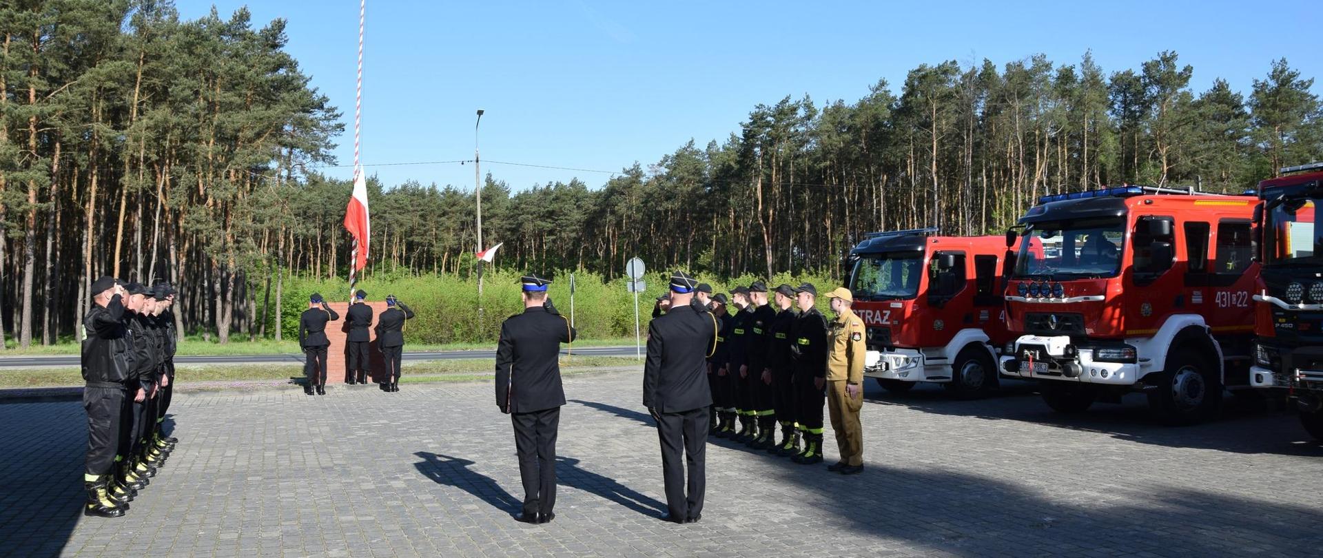 Zastępca Komendanta Powiatowego, Zastępca Dowódcy JRG oraz funkcjonariusze uczestniczący w uroczystej zbiórce stoją na baczność. Poczet flagowy wciąga flagę na maszt.