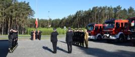 Zastępca Komendanta Powiatowego, Zastępca Dowódcy JRG oraz funkcjonariusze uczestniczący w uroczystej zbiórce stoją na baczność. Poczet flagowy wciąga flagę na maszt.