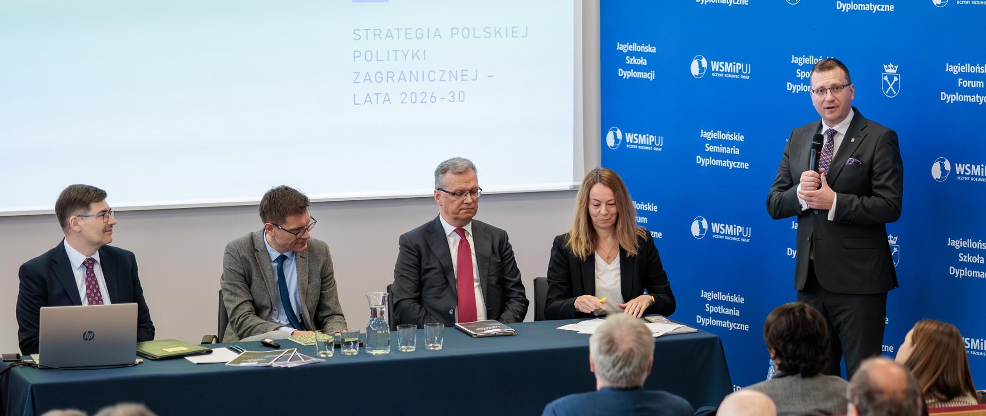 Na zdjęciu jest 5 osób, trzech mężczyzn i kobieta siedzą przy długim stole. Jeden mężczyzna stoi. Przemawiają do publiczności. Za nimi jest napis "Strategia Polskiej Polityki Zagranicznej 2026-2023". Obok jest niebieska ścianka z białym napisem "Jagiellońskie Spotkania Dyplomatyczne"