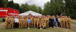 Szerokie ujęcie grupowe wykonane w plenerze bazy szkoleniowej w Kręsku. Zgromadzeni pozują uśmiechnięci w kilku rzędach na zielonej trawie. W tle po lewej stronie stoi duży, czerwony pojazd strażacki, a za całą grupą rozciąga się gęsty, zielony las pod jasnym, lekko zachmurzonym niebem. Zdjęcie przepełnione jest dumą, poczuciem wspólnoty, satysfakcją z udanej narady i integracją zespołu.