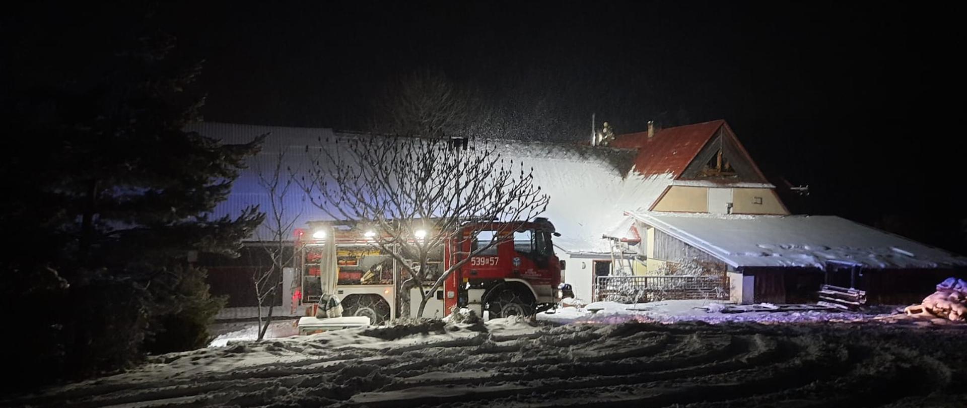 Zdjęcie przedstawia samochód pożarniczy na tle domu, w którym prowadzone są działania. 