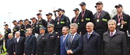 Na podium stoją mężczyźni, w górze trzymają puchary. Przed podium stoją goście honorowi w szeregu.