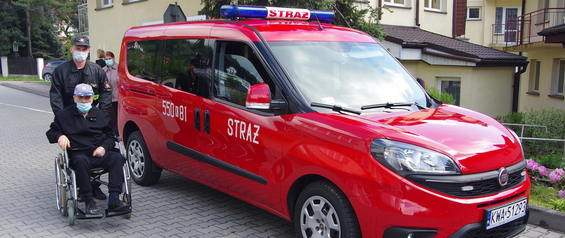 Widok z przodu. Strażak PSP w czarnym ubraniu koszarowym i maseczce ochronnej podjeżdża do czerwonego samochodu strażackiego z wózkiem inwalidzkim, na którym siedzi starszy mężczyzna w maseczce ochronnej. 