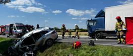 Zdjęcie przedstawia wypadek dwóch samochodów. Samochód osobowy z uszkodzonym przodem znajduje się w przydrożnym rowie. Samochód ciężarowy z uszkodzoną kabiną stoi na jezdni. Na jezdni stoją również samochody strażackie oraz karetka pogotowia ratunkowego. Przy parawanie stoją strażacy. 