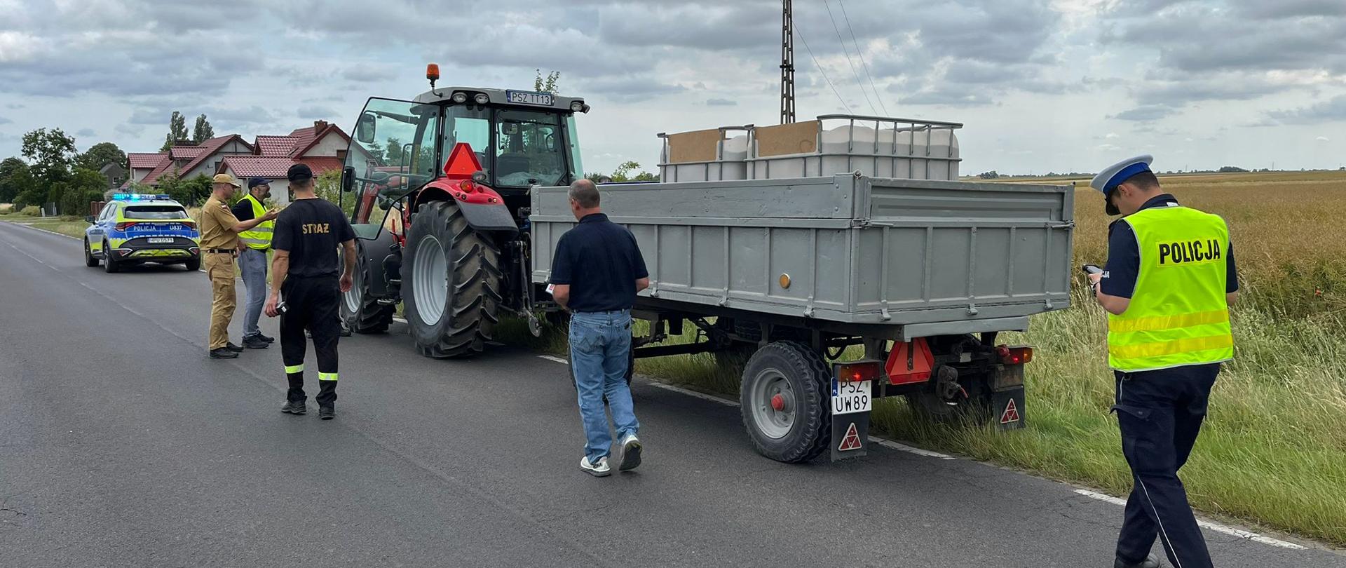 Ciągnik rolniczy z przyczepą na drodze, w pobliżu strażak i policjant
