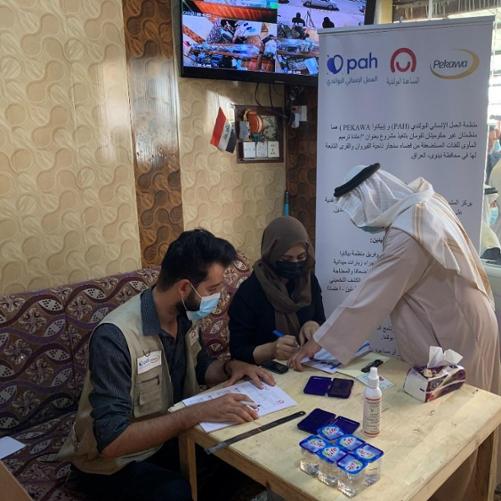 Kobieta i mężczyzna wypełniają dokumenty siedząc przy stole, a trzecia osoba zostawia odcisk kciuka na jednej z kartek. W tle rollup z logo Polskiej pomocy, PAH i Pekawa