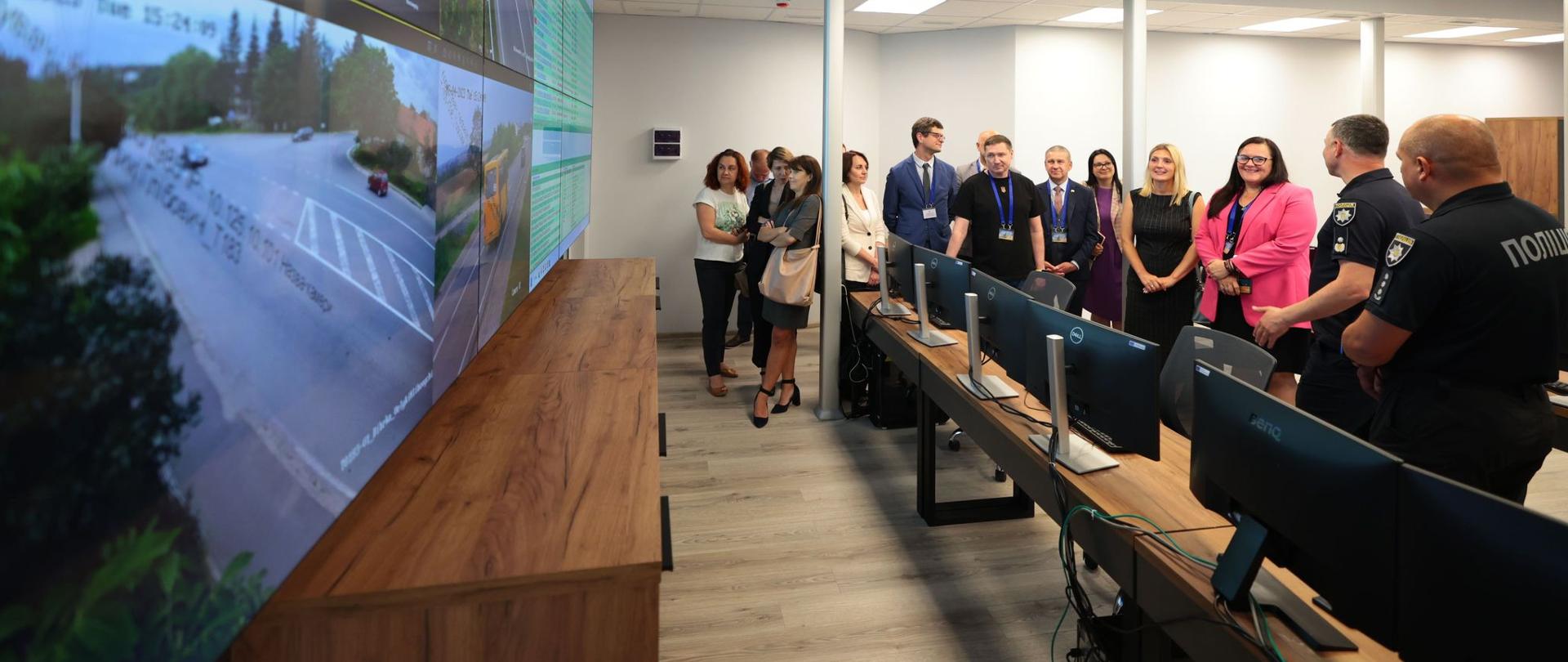 Grupa osób w pomieszczeniu. Przed nimi monitory na stołach, na ścianie ekrany,