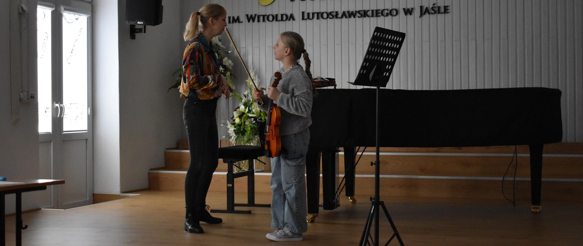 Dziewczynka w niebieskich spodniach i szarym swetrze w lewej ręce trzymająca skrzypce w prawej ręce do góry uniesione smyczek patrzy na kobietę o blond włosach w kolorowej bluzce i czarnych spodniach. Za nimi stoi czarny fortepian. W tle jasna ściana z logo szkoły i napisem Państwowa Szkoła Muzyczna I st. im Witolda Lutosławskiego w Jaśle..
Z prawej strony widać godło państwowe
