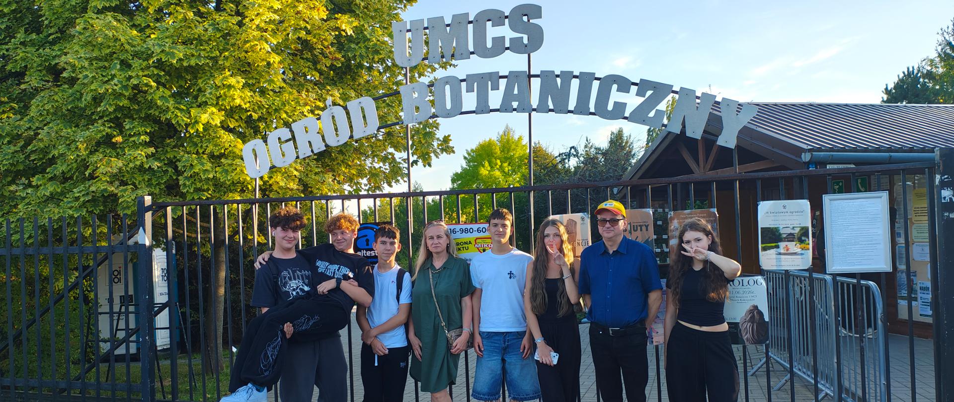 Grupa ośmiu osób stoi przed czarną bramą. Nad nimi znajduje się napis: UMSC OGRÓD BOTANICZNY. Pierwszy chłopak po lewej stronie trzyma kolegę na rękach. Dziewczęta pierwsza i trzecia od prawej pokazują dwa palce. Wszyscy się uśmiechają. Po lewej stronie z tyłu znajduje się zielone drzewo, a po prawej brązowy budynek.