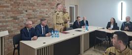 Odprawa roczna podsumowującą stan bezpieczeństwa ochrony przeciwpożarowej na terenie powiatu kolneńskiego za 2025 rok - zastępca podlaskiego komendanta wojewódzkiego PSP i komendant powiatowy PSP w Kolnie stoją przy stołach, przy których siedzą zaproszeni na uroczystość goście