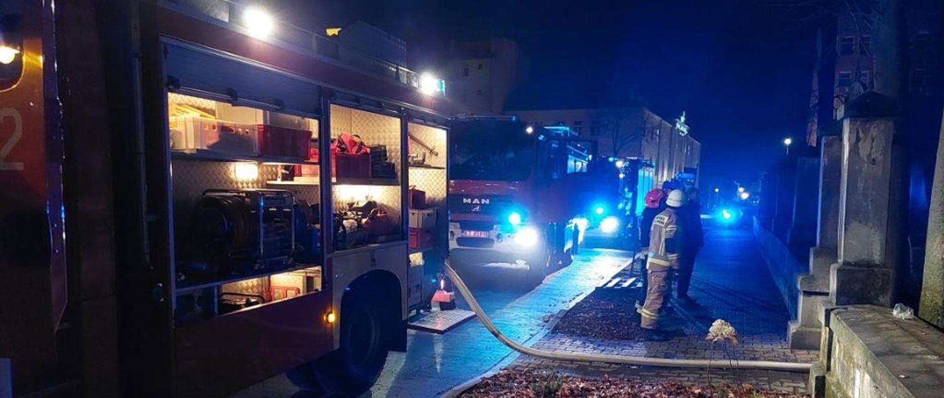 Na zdjęciu widać 3 samochody ratowniczo – gaśnicze podczas działań przy szpitalu im. E. Szczeklika oraz dwóch strażaków stojących obok linii wężowej.