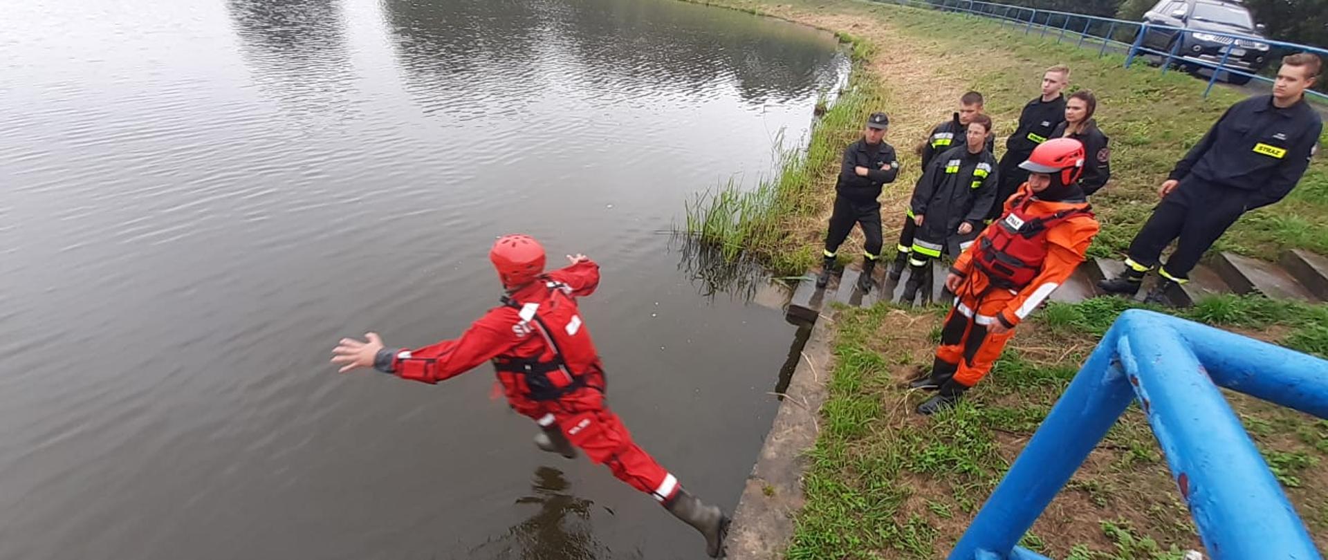 Strażacy w suchych skafandrach koloru czerwonego ćwiczą skoki do wody.