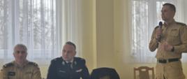 Na zdjęciu widoczny komendant powiatowy Państwowej Straży Pożarnej podczas przemówienia na naradzie operacyjnej dla naczelników OSP i komendantów gminnych ZOSP RP z terenu powiatu gorlickiego. Komendant ubrany w ubranie służbowe koloru pisakowego przemawia obok przy stole siedzą zastępca komendanta powiatowego w Gorlicach ubrani w ubrania służbowe koloru pisakowego, prezes powiatowy Związku Ochotniczych Straży Pożarnych Rzeczypospolitej Polskiej w Gorlicach ubrany w ubranie wyjściowe koloru granatowego.