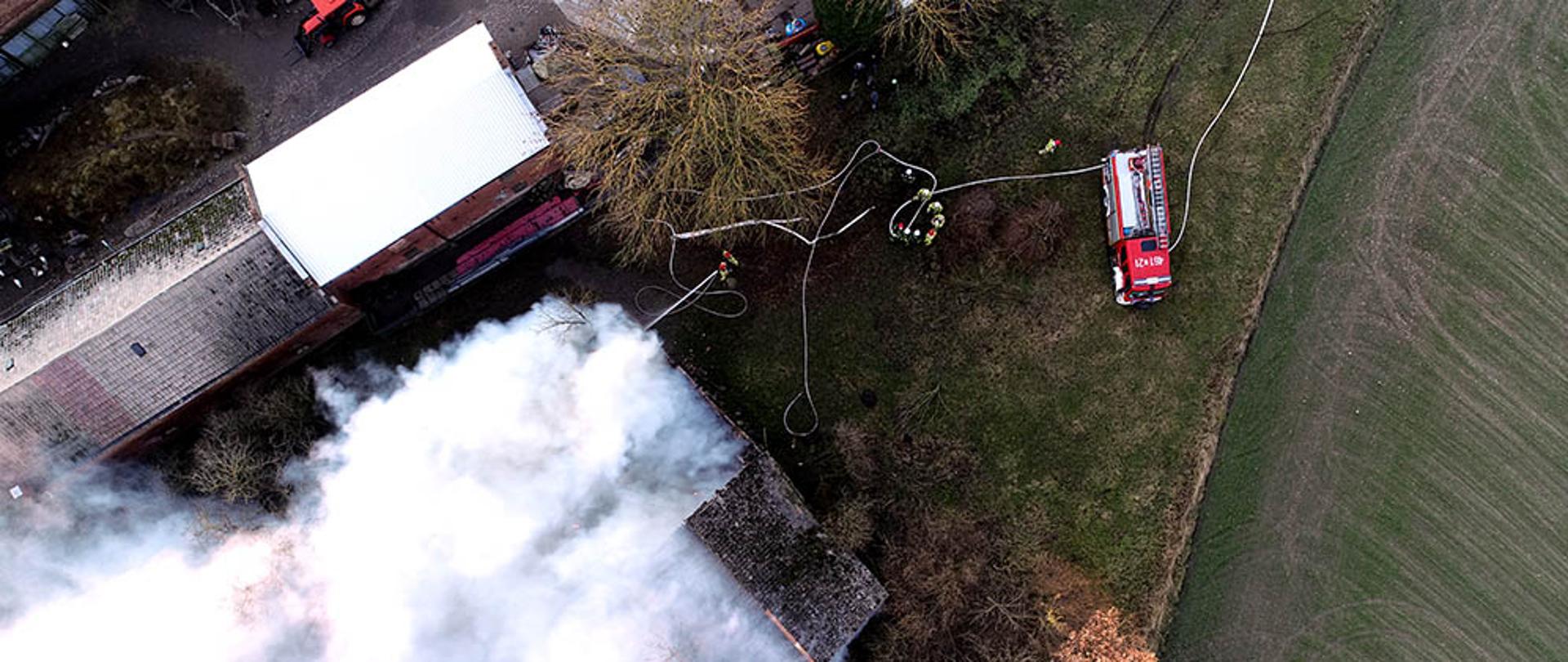 Zdjęcie z lotu ptaka przedstawia budynek objęty pożarem oraz działania straży pożarnej.