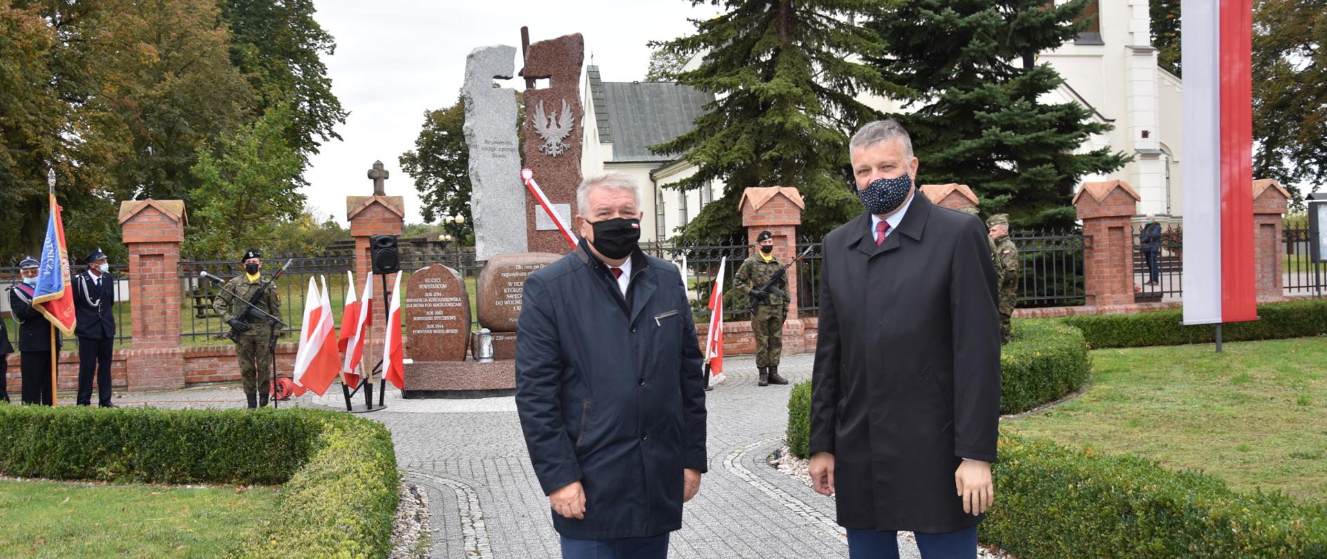 Elegancko ubrani mężczyźni stoją na tle szaro brązowego pomnika. W tle widać żołnierzy oraz kościół. 