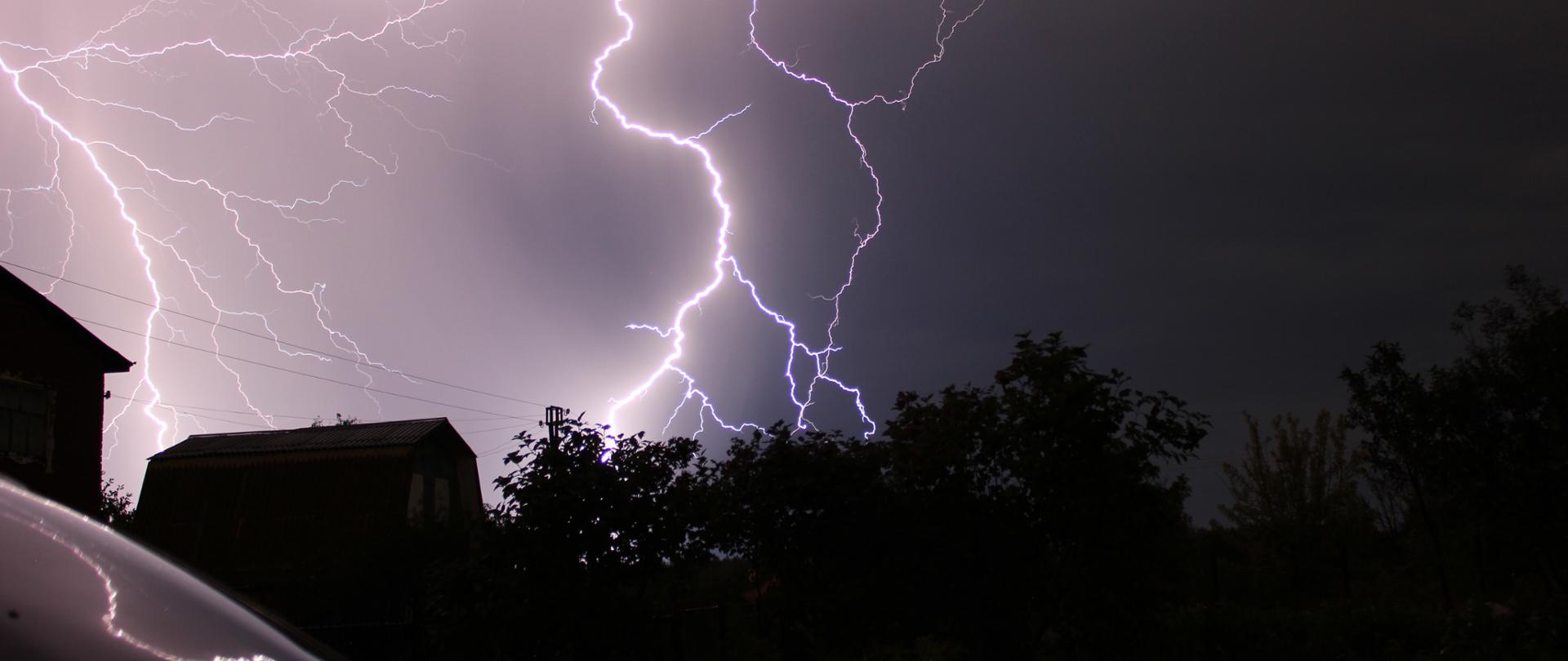Zdjęcie przedstawia burzę nocą. Widoczna jest duża błyskawica nad domami i koronami drzew.