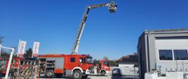 Samochód pożarniczy z otwartymi skrytkami przed zakładem produkcji okien. Z tyłu rozstawiony podnośnik strażacki podaje wodę na dach zakładu. Po lewej flagi z napisem STOLBUD okna & drzwi.