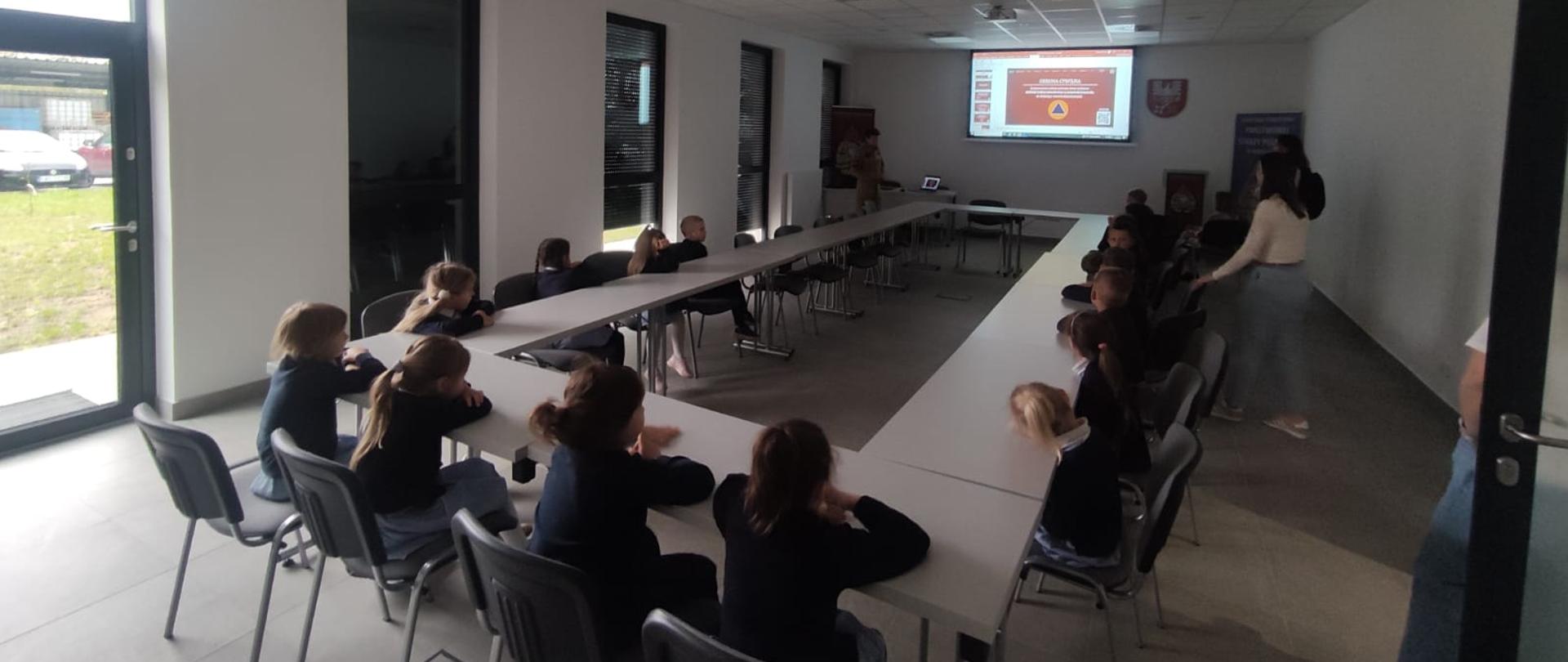 Na pierwszym planie widać dzieci słuchające prelekcji prowadzonej przez znajdującego się na drugim planie strażaka. W tle widoczny jest międzynarodowy znak obrony cywilnej. 