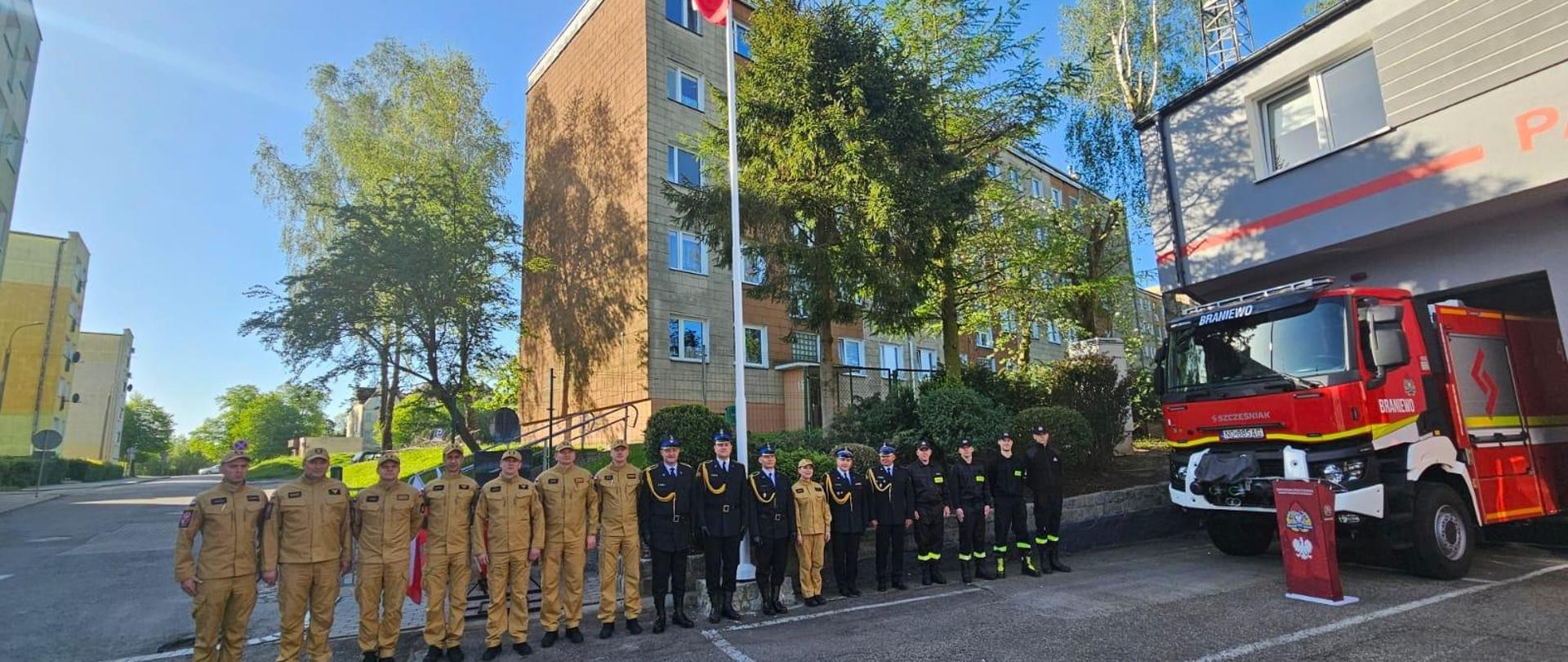 Na maszcie flaga Polski. Strażacy ustawieni obok siebie w mundurach. Z prawej strony samochód strażacki. Słonecznie.