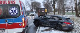 Uszkodzony samochód osobowy po zderzeniu z busem stojący w przydrożnym rowie, przód wystaje na jezdnię. Bok od strony kierowcy zniszczony. Obok stoi karetka pogotowia ratunkowego a w tle ambulans wojskowy.