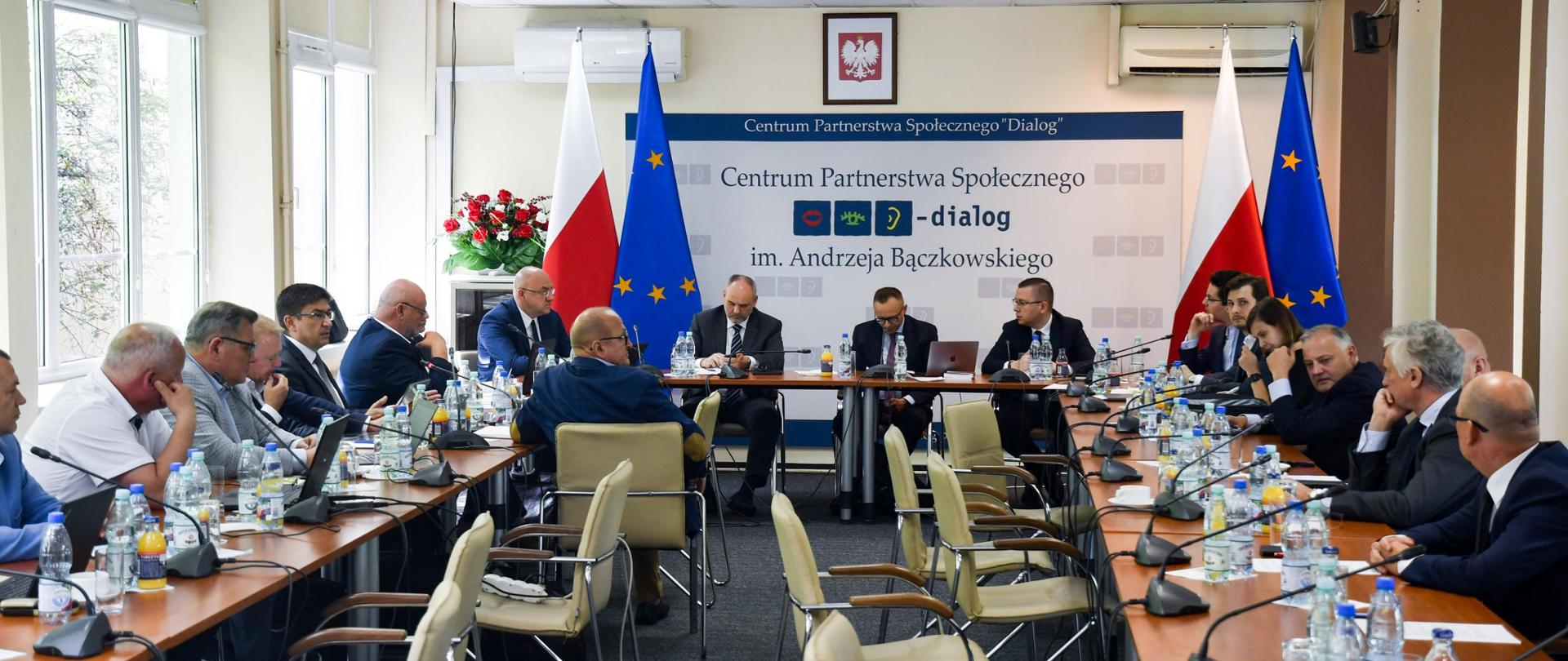 Sala konferencyjna. Przy długich stołach siedzą uczestnicy rozmów. U szczytu stołu siedzi wiceminister Artur Sasin. W tle ścianka Centrum Partnerstwa Społecznego Dialog, oraz flagi polskie i unijne.