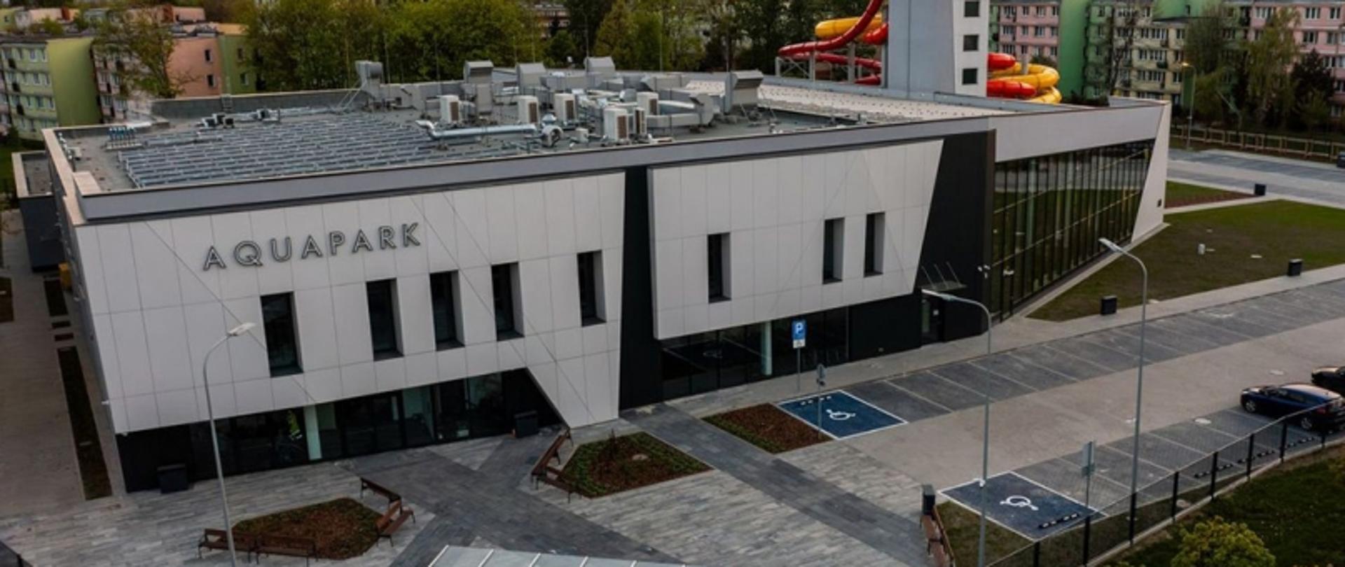 Budynek aquaparku z płaskim dachem i urządzeniami technicznymi, przed nim parking z miejscami dla niepełnosprawnych; w tle zjeżdżalnie i bloki mieszkalne.