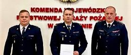 Wspólne zdjęcie en face stojących obok siebie od lewej bryg. Grzegorza Słoty, mł. bryg. Tomasza Samborskiego i st. bryg. Jarosława Tomczyka na tle godła PSP