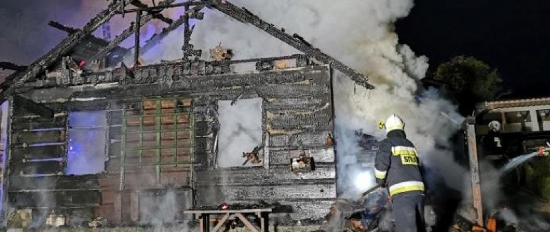 Zdjęcie przedstawia całkowicie spaloną zewnętrzną ścianę drewnianego budynku. Widoczne są fragmenty spalonego dachu w postaci mocno zwęglonych krokwi tworzących przedtem jego konstrukcje. Ponad dachem i przez uszkodzone w wyniku pożaru okna budynku widać unoszące się kłęby gęstego białego dymu. Obok budynku, w prawym jego rogu uchwycono postać strażaka ubranego w ubranie specjalne koloru granatowego w odblaskowymi popielato seledynowymi paskami na nogawkach spodni oraz rękawach i w dolnej i środkowej części kurtki, z białym kaskiem na głowie. Strażak ten w pozycji pół pochylonej zajmuje się dogaszaniem sterty spalonych rzeczy wyniesionych prawdopodobnie z wewnątrz objętych pożarem pomieszczeń. Przed spalonym budynkiem, pod oknem stoi nadpalona drewniana ławka.