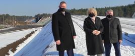 Dwóch mężczyzn i jedna kobieta stojących obok siebie na terenie inwestycji PKP. Ubrani elegancko. Stoją na budowanym wiadukcie pokrytym śniegiem.