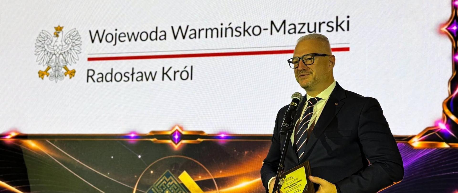 Mężczyzna w garniturze, wojewoda, stoi na tle wyświetlanego na prezentacji loga Wojewoda Warmińsko-Mazurski. Wojewoda przemawia przez mikrofon, uśmiecha się. 