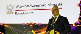 Mężczyzna w garniturze, wojewoda, stoi na tle wyświetlanego na prezentacji loga Wojewoda Warmińsko-Mazurski. Wojewoda przemawia przez mikrofon, uśmiecha się. 