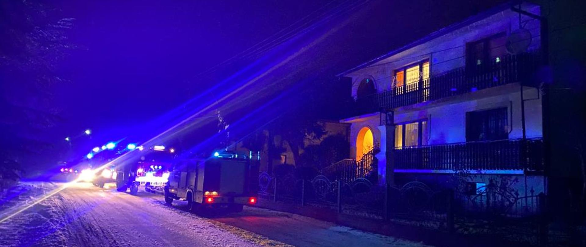 Na zdjęciu przedstawione są trzy samochody ratwoniczo-gaśnicze, które znajdują się na poboczu drogi obok budynku mieszkalnego. Samochody posiadają włączoną alarmową sygnalizację świetlną oraz włączone światła mijania. Zdjęcie zrobione jest w porze nocnej. W budynku mieszkalnym w kilku pomieszczeniach świeci się światło.