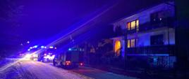Na zdjęciu przedstawione są trzy samochody ratwoniczo-gaśnicze, które znajdują się na poboczu drogi obok budynku mieszkalnego. Samochody posiadają włączoną alarmową sygnalizację świetlną oraz włączone światła mijania. Zdjęcie zrobione jest w porze nocnej. W budynku mieszkalnym w kilku pomieszczeniach świeci się światło.