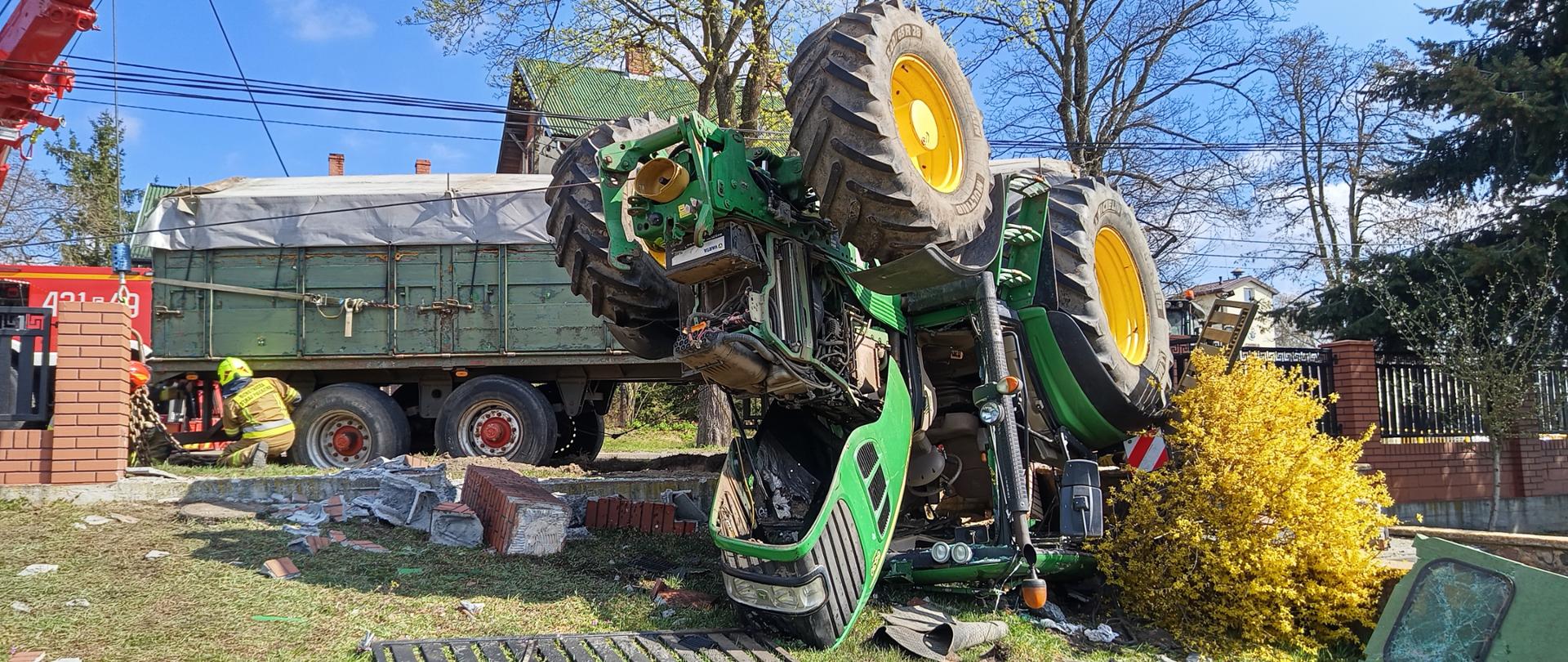 Fotografia przedstawia ciągnik leżący do góry kołami, za ciągnikiem widoczne zniszczone murowane słupki ogrodzenia, na dalszym planie przyczepa stojąca na kołach oraz pracujący strażak.