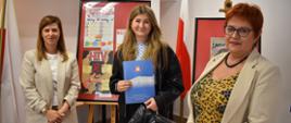 Trzy osoby stoją obok siebie w pomieszczeniu, na tle sztalug z oprawionymi plakatami edukacyjnymi dotyczącymi HIV/AIDS. Osoba znajdująca się pośrodku trzyma niebieską teczkę z logo oraz torbę z upominkami, na której widoczny jest napis „Podlaski Urząd Wojewódzki w Białymstoku”. Po obu stronach stoją dwie osoby w jasnych marynarkach. W tle znajdują się biało-czerwone flagi oraz plakaty na sztalugach. 