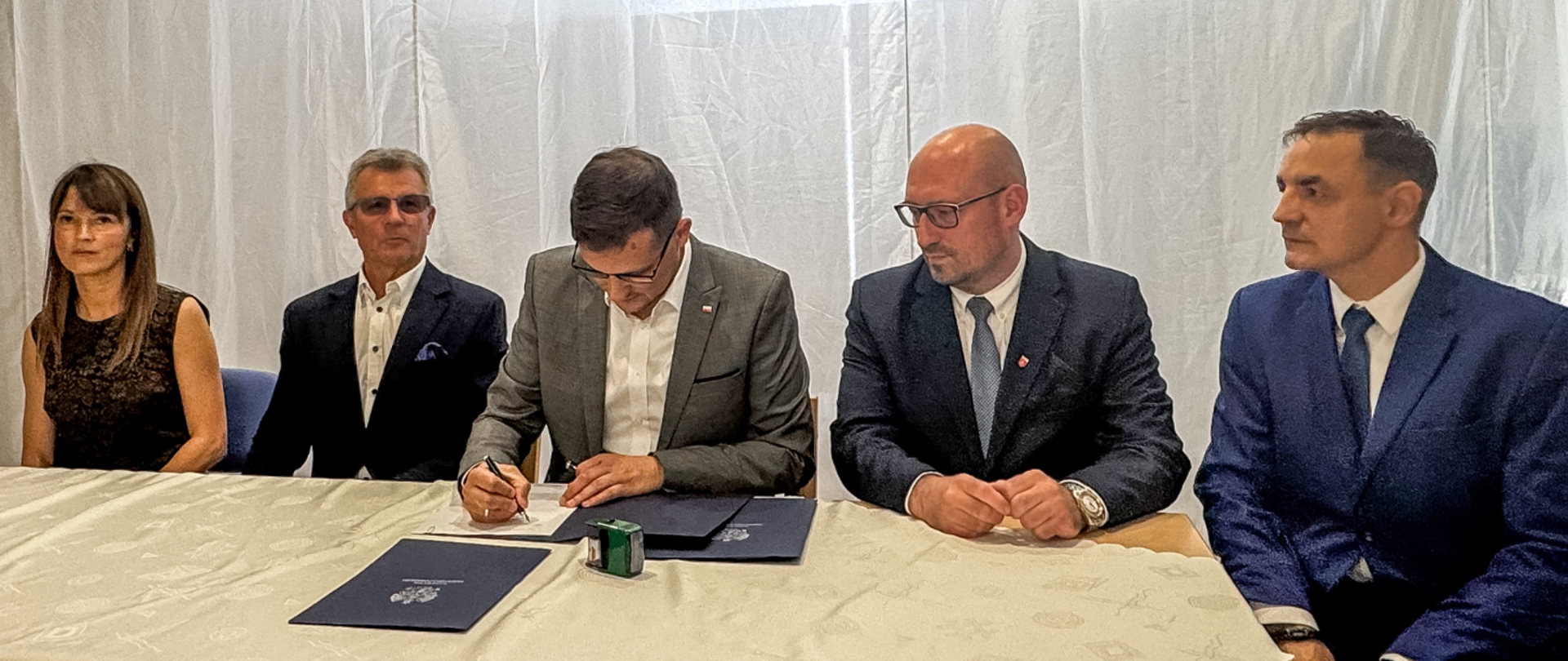 Sześć osób siedzi przy stole konferencyjnym. Wojewoda podpisuje dokument, a pozostali obserwują przebieg spotkania. Na stole znajdują się teczki z godłem Rzeczypospolitej Polskiej oraz pieczątka.