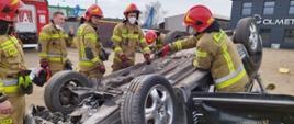 Na zdjęciu samochód osobowy leży na dachu. Strażacy w ubraniach specjalnych stoją wokół niego.