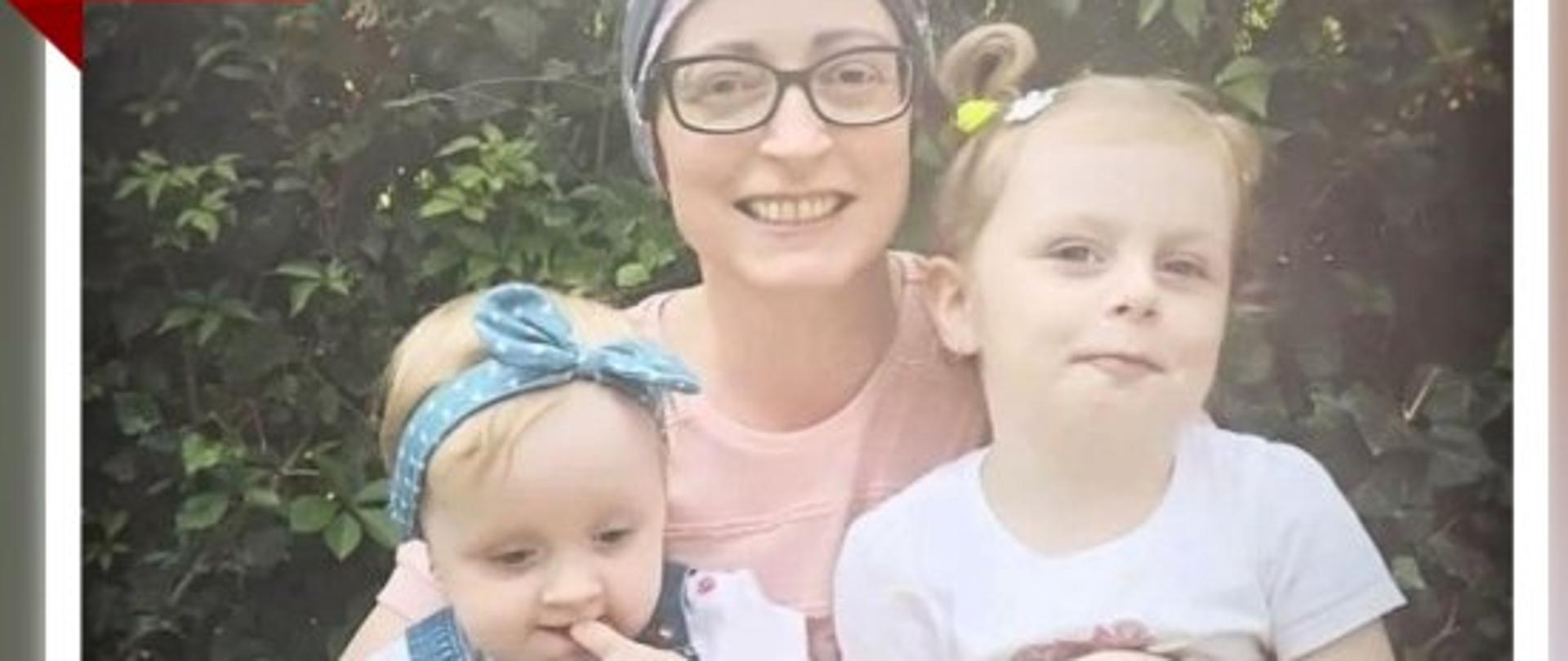 zdjęcie przedstawia uśmiechniętą kobietę z chustą na głowie, która obejmuje dwie małe dziewczynki. w tle 