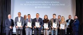 Zdjęcie przedstawia jedenaście osób stojących na scenie. Za osobami stojącymi na scenie wyświetlony jest napis "Zachodniopomorska Komisja ds. Bezpieczeństwa Pracy w Rolnictwie".