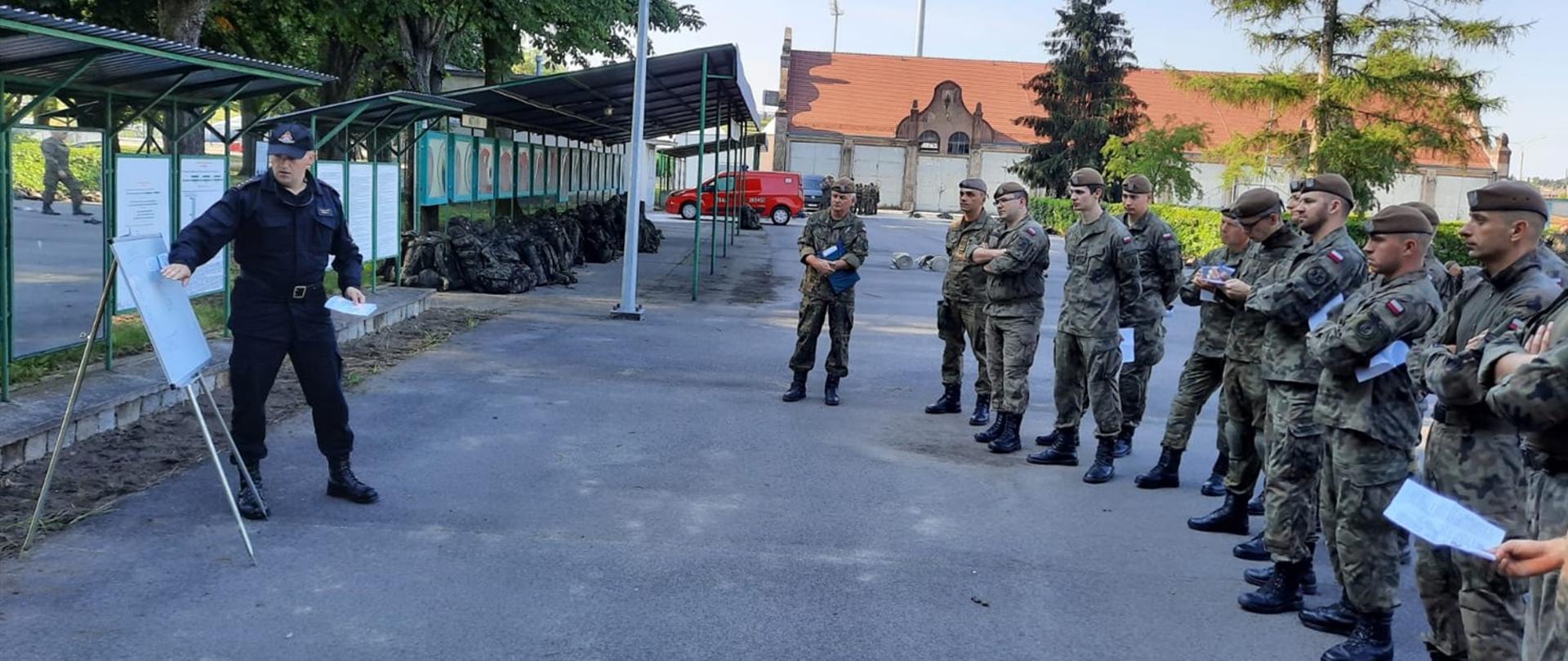 Na zdjęciu widoczny funkcjonariusz Państwowej Straży Pożarnej oraz grupa umundurowanych żołnierzy Wojsk Obrony Terytorialnej. W tle widoczne budynki jednostki wojskowej.