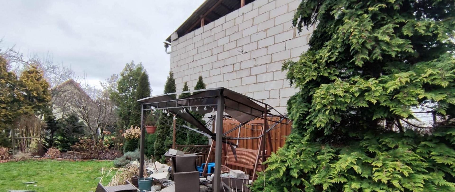 Wiatr uszkodził część szczytową budynku gospodarczego. Elementy budowlane runęły na aluminiową altankę ogrodową zlokalizowaną na sąsiedniej posesji. Altanka i jej wyposażenie została całkowicie uszkodzona. Na trawniku leżą elementy części szczytowej oraz uszkodzonej altanki