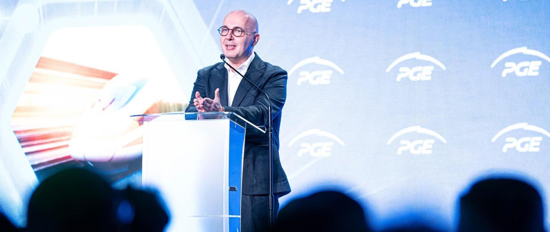 Wiceminister Robert Kropiwnicki stoi za mównicą. W tle ekran z logotypami PGE. Ujęcie zza słuchaczy. 