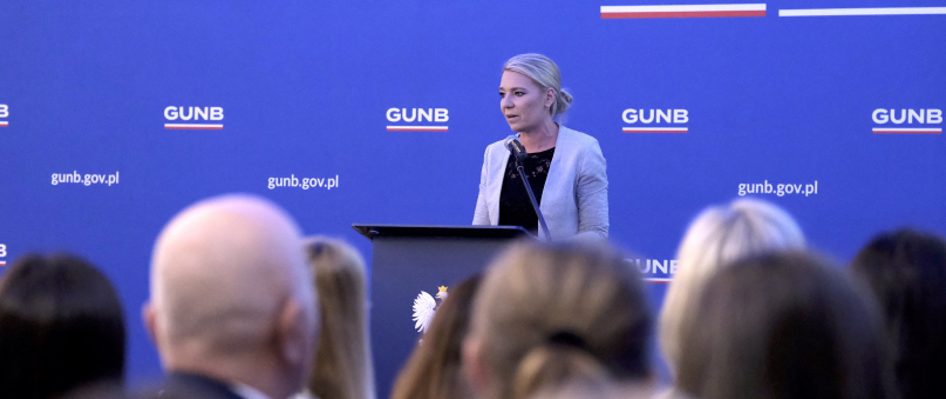 Osoba przemawiająca z mównicy na tle ścianki GUNB z logo i adresem gunb.gov.pl.