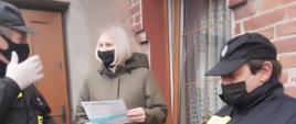 Zdjęcie przedstawia dwóch strażaków OSP, którzy wręczyli kobiecie ulotkę dotyczącą szczepień przeciwko COVID-19. Zdjęcie wykonane jest na schodach przed wejściem do budynku mieszkalnego. 