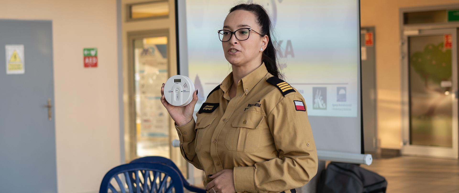 Kobieta w brązowym mundurze z naszywkami, w okularach i z długimi, ciemnymi włosami, stoi i trzyma w dłoniach czujkę dymu. W tle widać rzutnik z prezentacją oraz krzesła. Kobieta stoi na tle dużej sali.