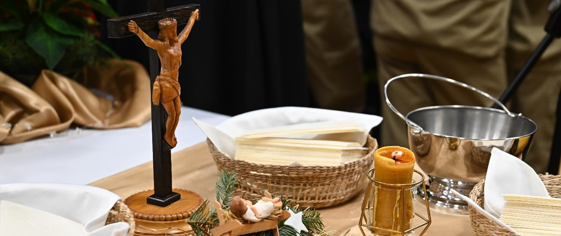 Na stoliku stoi krzyż, obok sianko ze żłobkiem i Panem Jezusem w koszykach znajdują się białe opłatki. Na stole stoją też dwie zapalone świece