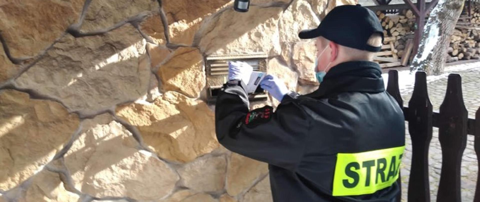 Na zdjęciu strażak w umundurowaniu koszarowym wrzucający ulotkę do skrzynki pocztowej dot. szczepień na covid-19